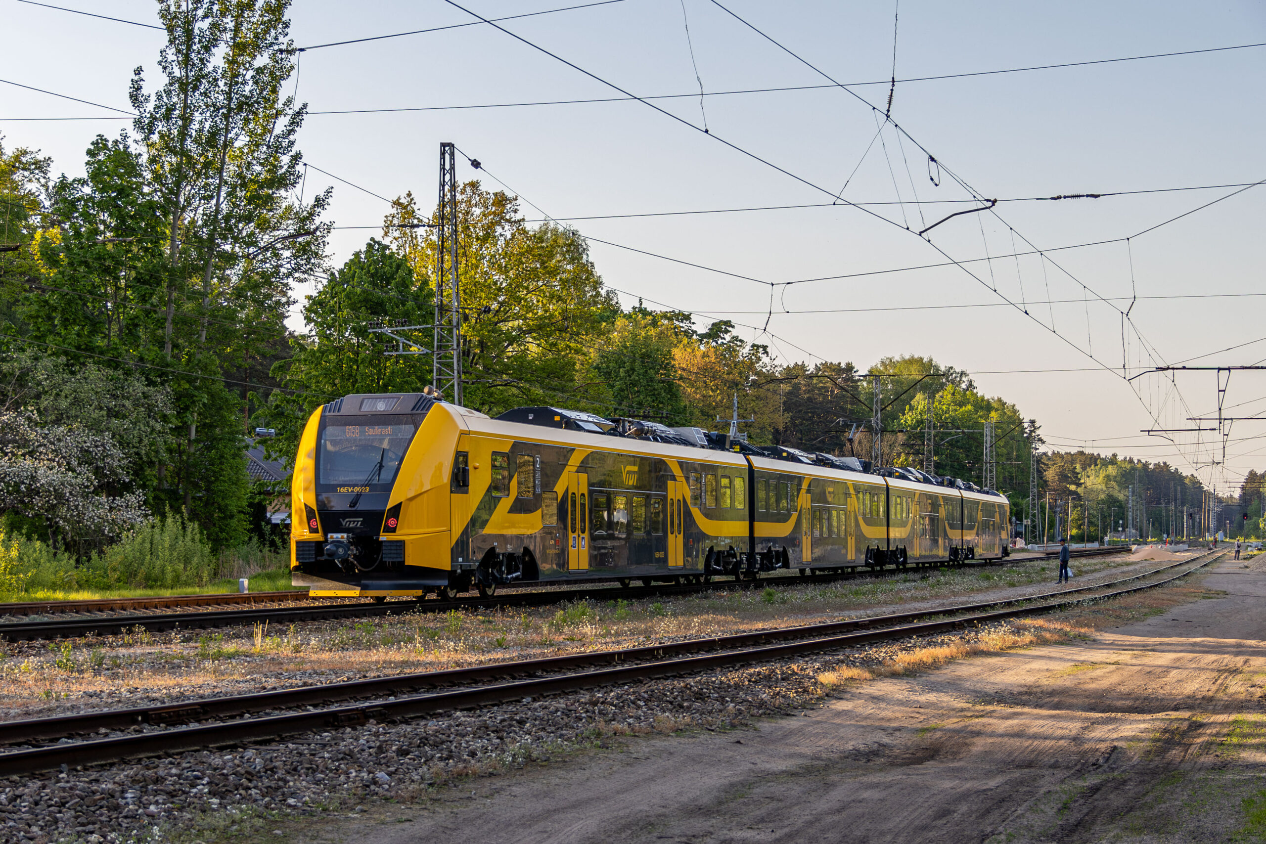 The image illustrates Škoda 16Ev electric passenger train with livery of Latvian national passenger rail carrier brand Vivi departing a station in Latvia enroute to Saulkrasti